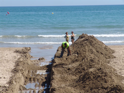 Uns operaris fent la rasa a la platja d'Adarró mentre unes banyistes passegen per la sorra.