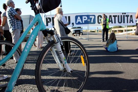 Part d'una bicicleta en primer pla amb una nena asseguda a l'asfalt de l'N-340 mentre el portaveu del moviment veïnal, Llorenç Navarro, parla . ACN