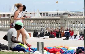 Pla general d'un manter al Passeig de la Ribera de Sitges, assegut al terra mentre dues turistes passegen pel costat. ACN