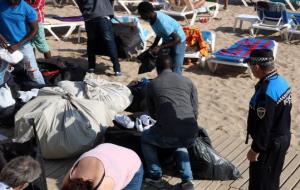 Pla obert d'un grup de venedors del top manta recollint les seves pertinences a la platja de Sitges sota l'observació d'un agent de la Policia Local. 