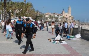 Pla general de dos agents de la Policia Local de Sitges patrullant al passeig de la Ribera, amb la Punta al fons, mentre un manter recull. ACN