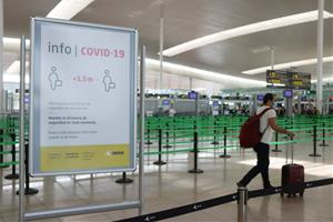 Un cartell informatiu de la covid-19 i d'un passatger arrossegant una maleta amb el control de seguretat de la T1 de l'Aeroport del Prat. ACN