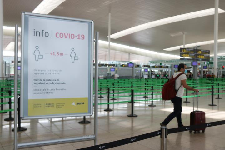 Un cartell informatiu de la covid-19 i d'un passatger arrossegant una maleta amb el control de seguretat de la T1 de l'Aeroport del Prat. ACN