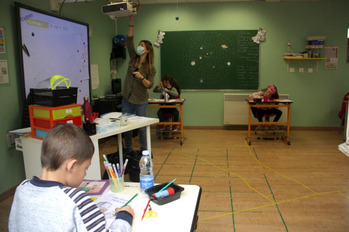 Una professora amb mascareta i utilitzant el projector de l'aula i un grup d'alumnes distanciats entre ells. ACN / Albert Lijarcio