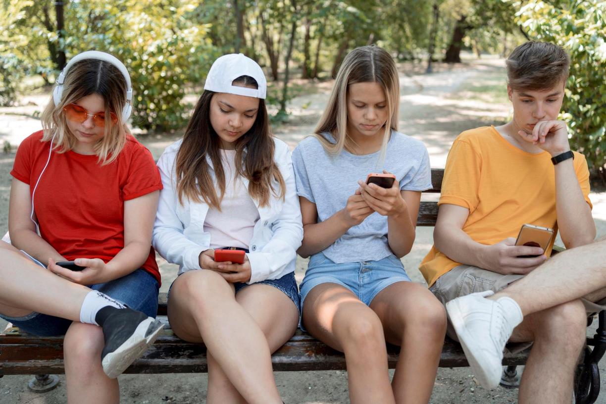 Adolescents asseguts en un banc al parc, centrats en els seus telèfons mòbils. Freepik
